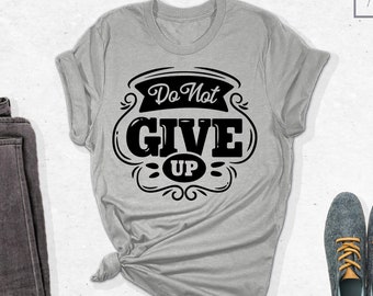 May include: A gray t-shirt with the text "Do Not Give Up" in a black vintage-style font. The shirt is folded and displayed with a pair of dark gray jeans and two pairs of blue shoes with orange laces.