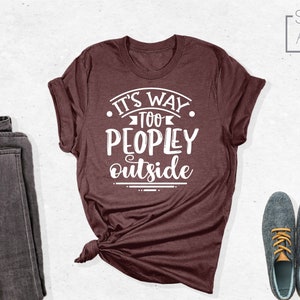 May include: A maroon t-shirt with the text "It's way too peopley outside" in white lettering. The shirt is folded and displayed on a white background with a pair of dark grey jeans and two dark blue shoes with tan laces.