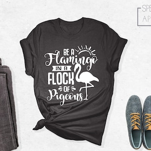 May include: A dark gray t-shirt with a white graphic that says "Be a Flamingo in a Flock of Pigeons". The graphic also includes a white flamingo silhouette.
