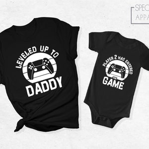 May include: Black t-shirt with white text that reads "Leveled Up To Daddy" and a video game controller graphic. A black baby onesie with white text that reads "Player 2 Has Entered The Game" and a video game controller graphic.
