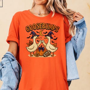 May include: Orange t-shirt with the word "GOOSEBUMPS" in orange and black lettering. The graphic features two cartoon ducks wearing witch hats, a pumpkin, a football, and a crescent moon. The shirt is a Halloween-themed design.