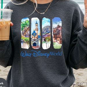 A woman wearing a gray sweatshirt with a graphic design of Disney World landmarks, including Cinderella Castle, the Eiffel Tower, and the Hollywood sign, along with a peace sign. She is holding a drink in a plastic cup with a straw.