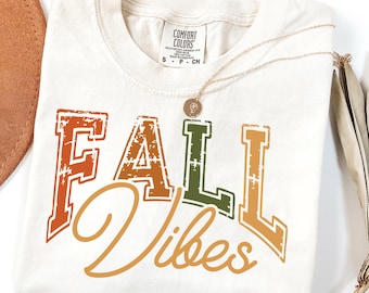 May include: Ivory-colored t-shirt with the words "FALL Vibes" in distressed orange, green, and gold lettering. A gold necklace and a brown hat are next to the shirt. The shirt is from Comfort Colors.