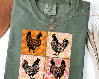 May include: A moss green t-shirt featuring a four-panel design with black rooster illustrations. Each rooster is filled with floral patterns and set against backgrounds of pumpkins and flowers. The shirt has a crew neck and short sleeves.