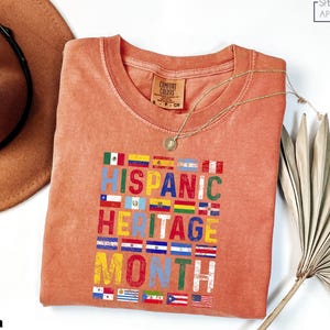 May include: A coral-colored t-shirt with the text "HISPANIC HERITAGE MONTH" in colorful letters, surrounded by flags. A gold necklace and a brown hat are also in the image. The shirt is made of a soft material.