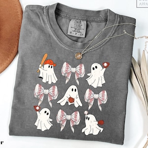 May include: A gray Comfort Colors t-shirt featuring a playful design of cartoon ghosts with baseball bats, gloves, and baseball-themed bows. The shirt has a crew neck and short sleeves. The word "Pepper" is printed at the bottom left.