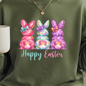 May include: Olive green sweatshirt featuring a colorful graphic of three Easter bunnies with bows and hearts. The words "Happy Easter" are written in a script font below the bunnies. The sweatshirt is a casual, festive garment.