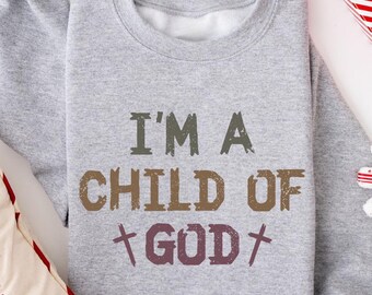 May include: A gray sweatshirt with the text "I'M A CHILD OF GOD" in brown, green, and purple. The sweatshirt is surrounded by Christmas decorations, including a pillow, a gift box, and a snowflake ornament.