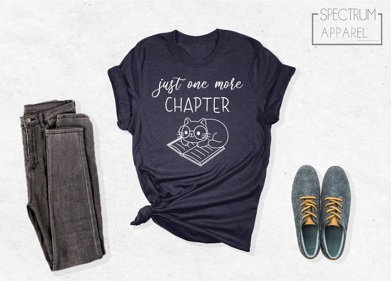 May include: A navy blue t-shirt with the text "just one more chapter" and a cartoon cat wearing glasses and reading a book. The shirt is folded and laid on a white surface with a pair of gray jeans and gray shoes.