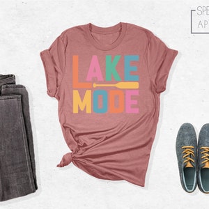 May include: A maroon t-shirt with the text "LAKE MODE" in colorful letters and a yellow paddle graphic. The shirt is folded and displayed on a white background.