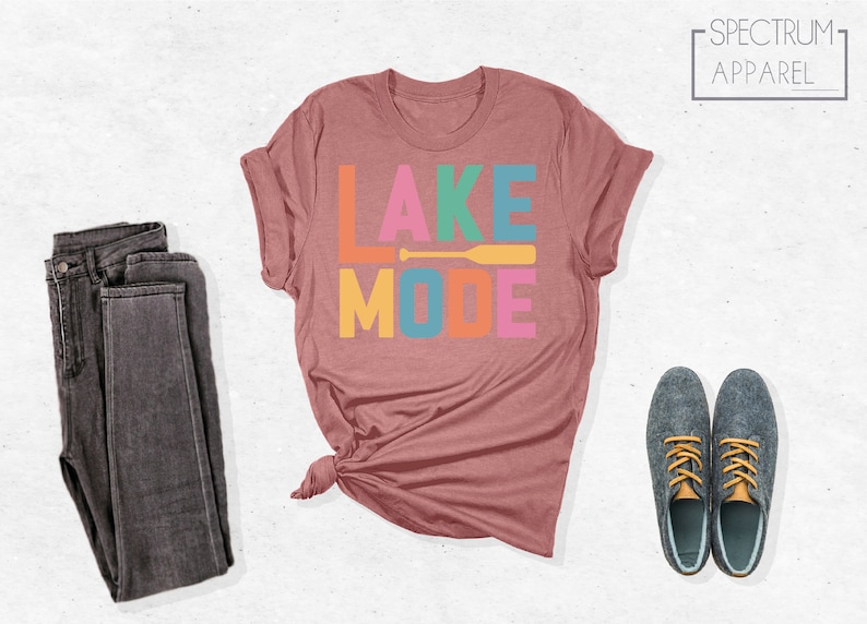 May include: A maroon t-shirt with the text "LAKE MODE" in colorful letters and a yellow paddle graphic. The shirt is folded and displayed on a white background.
