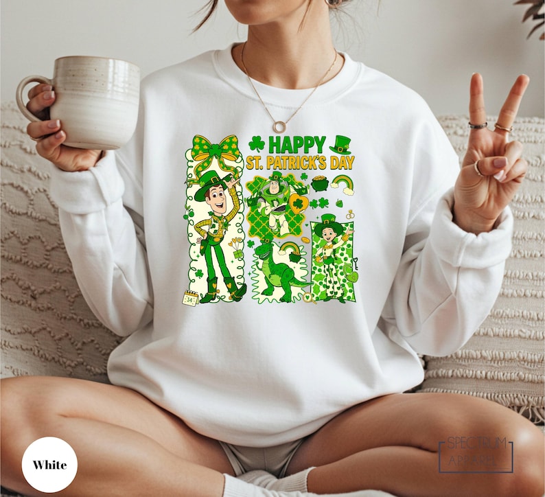 A woman wearing a white sweater with a graphic design of St. Patrick's Day characters and text, holding a mug and making a peace sign.
