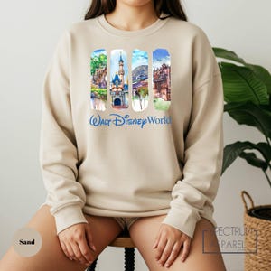 A woman wearing a beige sweater with a collage of Disney World images on it, sitting on a stool in front of a plant.
