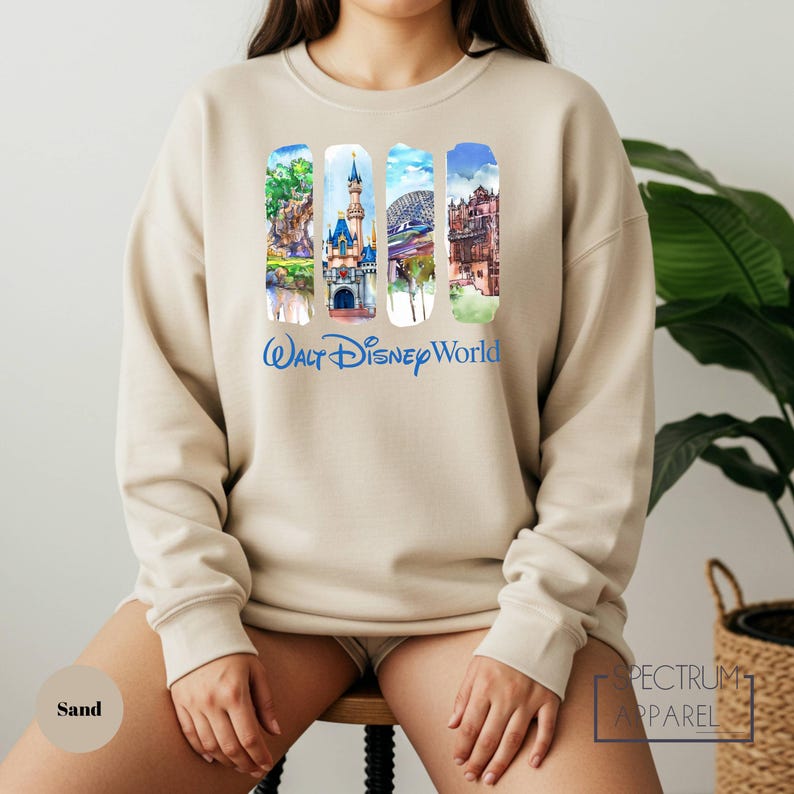 A woman wearing a beige sweater with a collage of Disney World images on it, sitting on a stool in front of a plant.