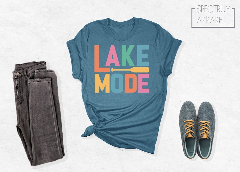 May include: A teal blue t-shirt with the text "LAKE MODE" in colorful block letters. A yellow paddle is included in the design. The text "SPECTRUM APPAREL" is in a white box in the upper right corner of the image.