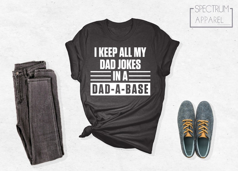 May include: A dark gray t-shirt with white text that reads "I keep all my dad jokes in a dad-a-base". The shirt is folded and tied in a knot. A pair of dark gray jeans and a pair of blue shoes are also pictured.