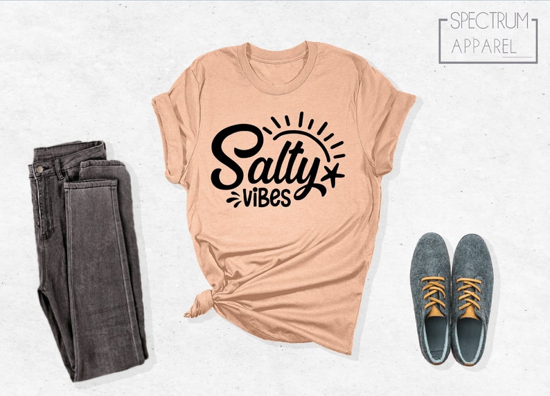 May include: A peach colored t-shirt with the text "Salty Vibes" in black lettering with a sun and a star graphic. The shirt is tied in a knot at the bottom. A pair of dark gray jeans and two gray shoes with yellow laces are also pictured.