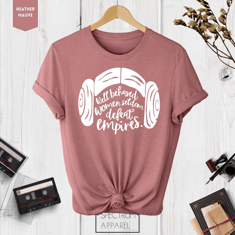 Puede incluir: Una camiseta de color malva jaspeado con un gr&aacute;fico blanco de la cabeza de una mujer con un mo&ntilde;o lateral y el texto "Well behaved women seldom defeat empires."