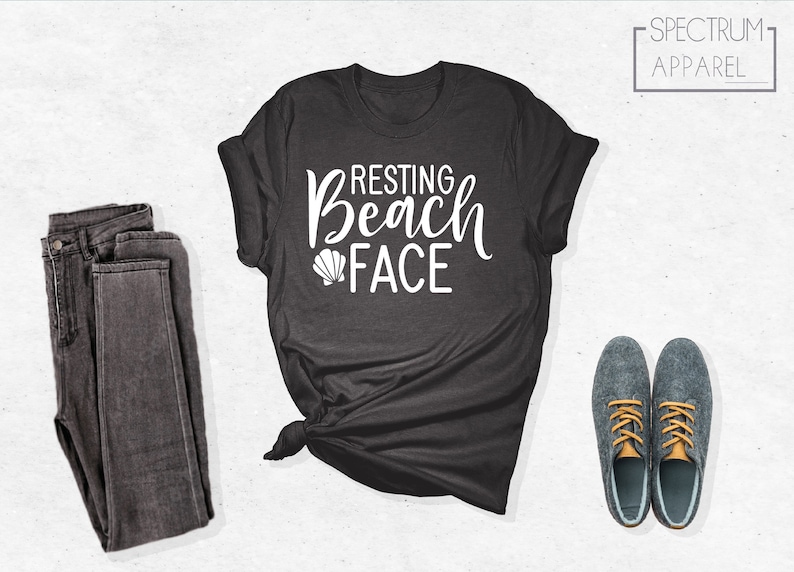 Może przedstawiać: Szary t-shirt z napisem "Resting Beach Face" w białym kolorze i małym nadrukiem muszelki. Koszulka jest zawiązana na dole.