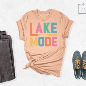 May include: A peach-colored t-shirt with the text "LAKE MODE" in rainbow colors. The text is stacked vertically with a small orange paddle graphic between the words.