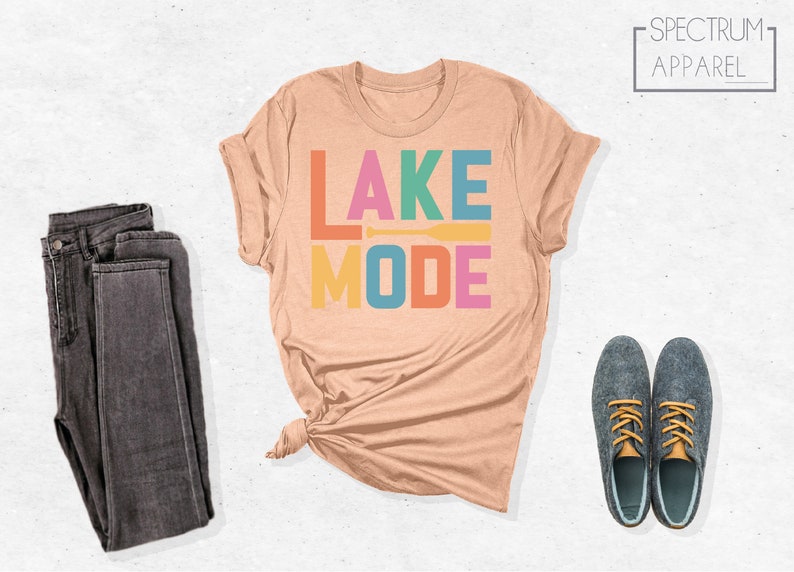 May include: A peach-colored t-shirt with the text "LAKE MODE" in rainbow colors. The text is stacked vertically with a small orange paddle graphic between the words.