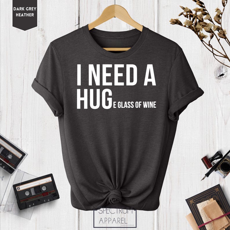 May include: Dark grey heather t-shirt with white text that reads "I need a hug and a glass of wine".