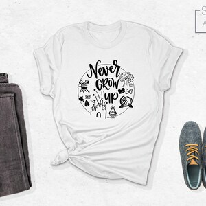 May include: White t-shirt with a black circle graphic featuring the text "Never Grow Up" and various Disney park icons. The shirt is folded and laid flat on a white surface.