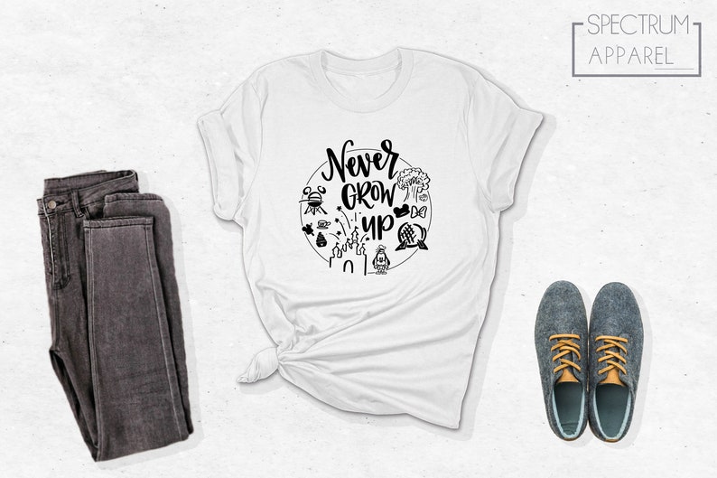 May include: White t-shirt with a black circle graphic featuring the text "Never Grow Up" and various Disney park icons. The shirt is folded and laid flat on a white surface.