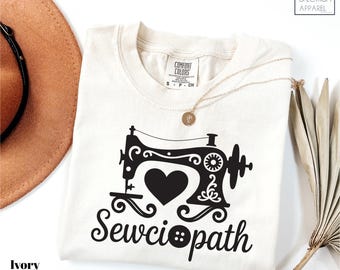May include: Ivory-colored t-shirt with a black sewing machine graphic and the word "Sewciopath". The design includes a heart and a button. The shirt is from Comfort Colors and Spectrum Apparel.