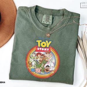 a green t-shirt with a colorful graphic of characters from the Toy Story movie, including Woody, Buzz Lightyear, and other beloved characters. The t-shirt is laid out on a white surface, with a brown hat and some leaves visible in the background.