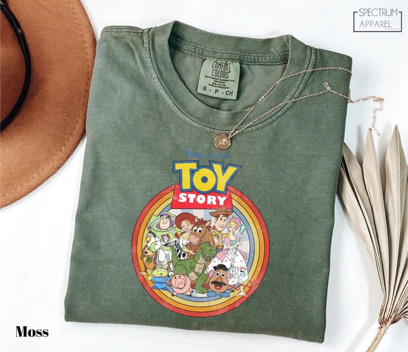 a green t-shirt with a colorful graphic of characters from the Toy Story movie, including Woody, Buzz Lightyear, and other beloved characters. The t-shirt is laid out on a white surface, with a brown hat and some leaves visible in the background.