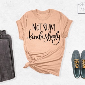 May include: A peach-colored t-shirt with the text "Not Slim Kinda Shady" in black lettering. The shirt is folded and displayed on a white background with a pair of dark gray jeans and two dark blue shoes with yellow laces.