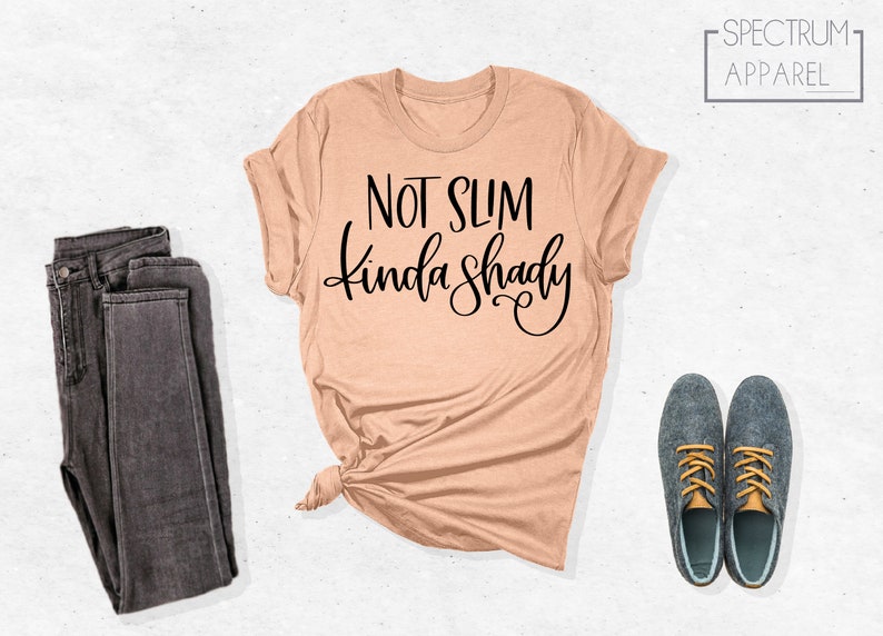 May include: A peach-colored t-shirt with the text "Not Slim Kinda Shady" in black lettering. The shirt is folded and displayed on a white background with a pair of dark gray jeans and two dark blue shoes with yellow laces.