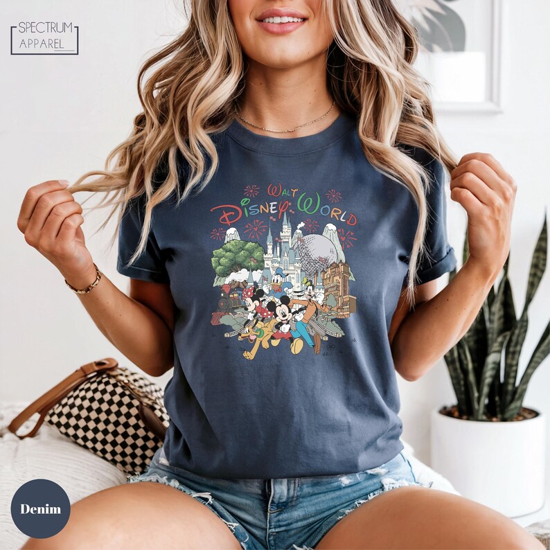 A woman wearing a blue t-shirt with a Disney World graphic and text, smiling and posing with her hands in the air.