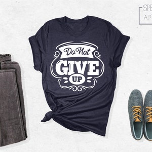 May include: A navy blue t-shirt with a white graphic that says "Do Not Give Up" in a vintage style font. The shirt is folded and laid flat on a white surface.