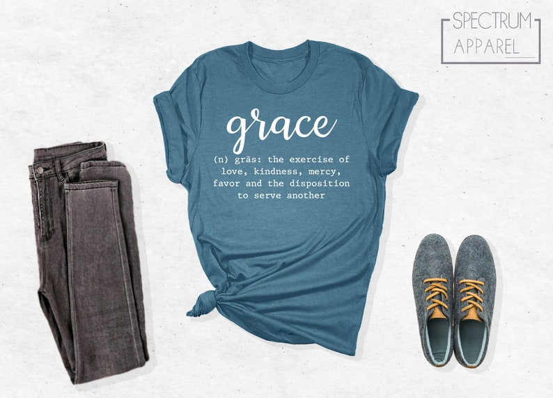 Op de afbeelding: Een tealblauw t-shirt met het woord "grace" in witte, cursieve letters. De definitie van genade staat eronder in witte letters: "(n) gr&auml;s: de uitoefening van liefde, vriendelijkheid, barmhartigheid, gunst en de bereidheid om een ander te dienen".