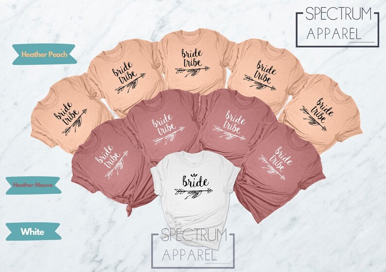 May include: Set of eight t-shirts in various shades of peach and mauve with the text "bride tribe" and an arrow graphic. One white t-shirt with the text "bride" and an arrow graphic. The shirts are arranged in a circle on a white marble surface.