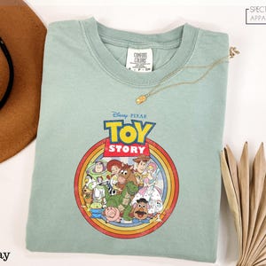 A green t-shirt with a Disney Pixar logo and characters from the Toy Story movie, along with a brown hat and a fan, are displayed on a white surface.