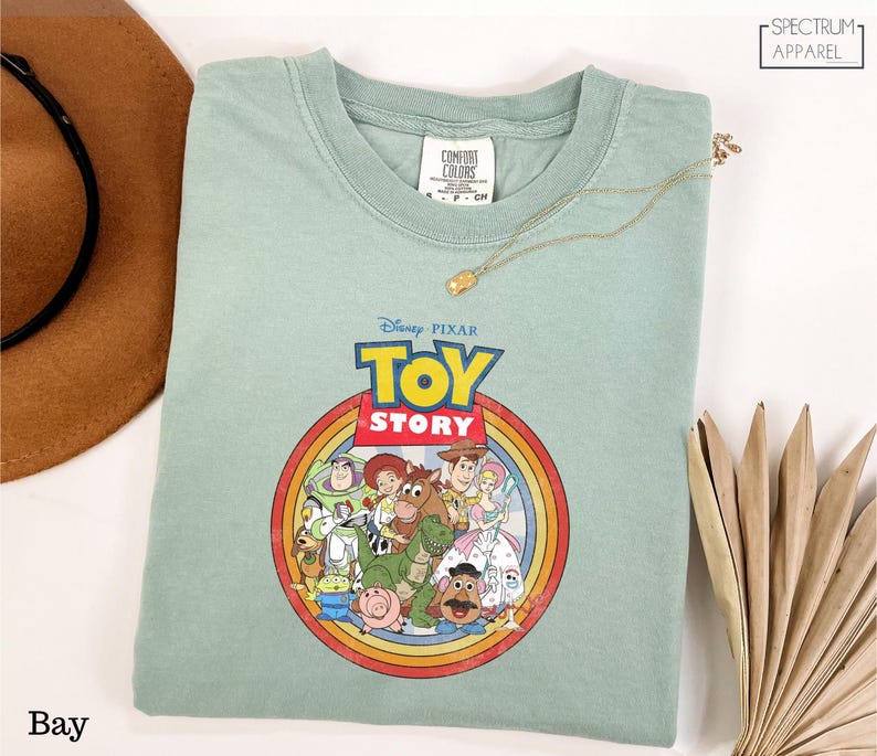 A green t-shirt with a Disney Pixar logo and characters from the Toy Story movie, along with a brown hat and a fan, are displayed on a white surface.