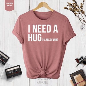 May include: A heather mauve t-shirt with the text "I need a hug and a glass of wine" printed in white.