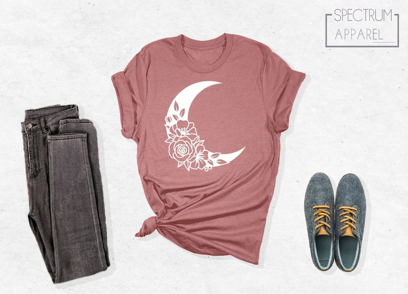 May include: A mauve t-shirt with a white crescent moon design featuring floral details. The t-shirt is folded and displayed on a white background with a pair of dark grey jeans and a pair of grey shoes with yellow laces.