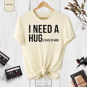 May include: A cream colored t-shirt with the text "I need a hug and a glass of wine" printed in black.
