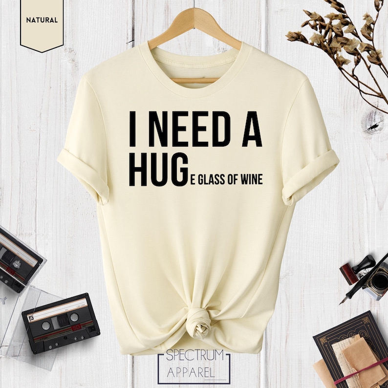 May include: A cream colored t-shirt with the text "I need a hug and a glass of wine" printed in black.