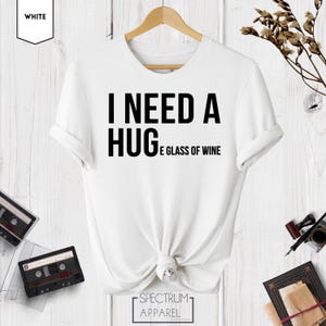 May include: White t-shirt with black text that reads "I need a hug and a glass of wine".