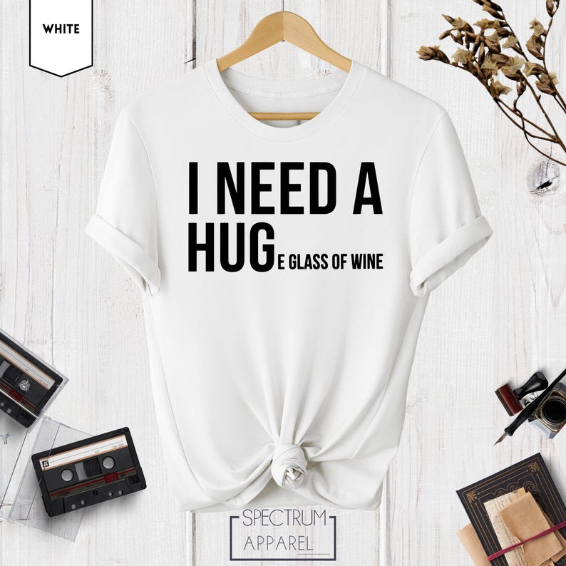 May include: White t-shirt with black text that reads "I need a hug and a glass of wine".