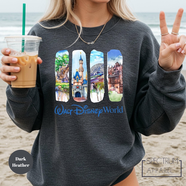 A woman wearing a gray sweatshirt with a graphic design of Disney World landmarks, including Cinderella Castle, the Eiffel Tower, and the Hollywood sign, along with a peace sign. She is holding a drink in a plastic cup with a straw.