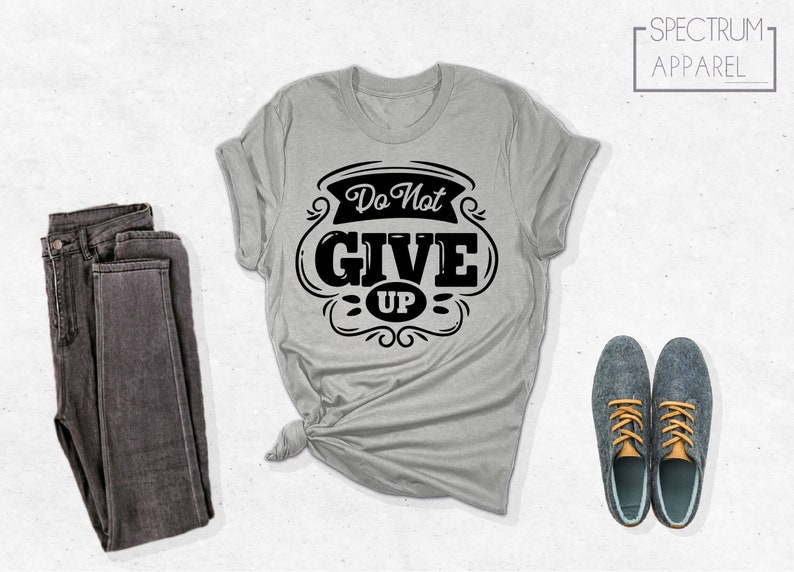 May include: A gray t-shirt with the text "Do Not Give Up" in a black vintage-style font. The shirt is folded and displayed with a pair of dark gray jeans and two pairs of blue shoes with orange laces.