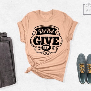 May include: A peach-colored t-shirt with a black graphic that says "Do Not Give Up" in a vintage style font. The shirt is folded and laid out on a white surface with a pair of dark gray jeans and two dark blue shoes with yellow laces.