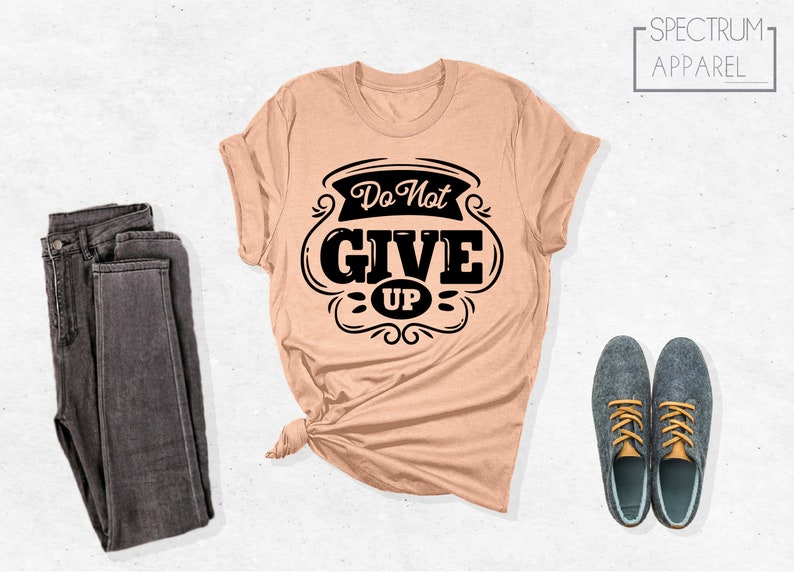 May include: A peach-colored t-shirt with a black graphic that says "Do Not Give Up" in a vintage style font. The shirt is folded and laid out on a white surface with a pair of dark gray jeans and two dark blue shoes with yellow laces.