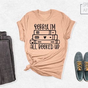 May include: A peach-colored t-shirt with a black graphic of a stack of books and the text "Sorry, I'm all booked up".